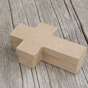 May include: A cross-shaped box made of light brown cardboard. The box is sitting on a weathered wooden surface. The cross design is a simple, geometric shape, with a lid that fits snugly over the base.