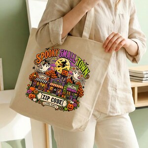 May include: A beige canvas tote bag featuring a colorful graphic print with the text SPOOKY SMALL TOWN, ZIP CODE, and TOWN NAME. The illustration depicts a Halloween-themed street scene with ghosts, a witch on a broom, pumpkins, and buildings.