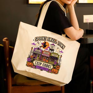 May include: A cream-colored canvas tote bag featuring a colorful Halloween-themed graphic. The design includes a witch on a broom, ghosts, pumpkins, and storefronts labeled Witch's Emporium, Pumpkin Bakery, and Curiosities, with text reading SPOOKY SMALL TOWN, [ZIP CODE], and [TOWN NAME].