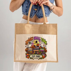 May include: A tan jute tote bag with a white front pocket featuring a colorful Halloween-themed graphic. The design includes a small town scene with pumpkins, ghosts, a witch on a broomstick, and the text SPOOKY SMALL TOWN and [ZIP CODE].