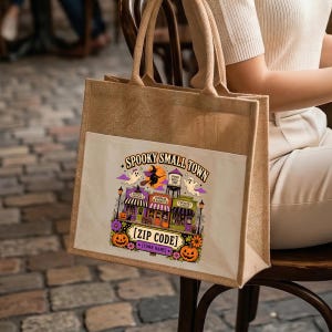 May include: A tan jute tote bag with two handles and a white front pocket featuring a colorful graphic of a spooky small town. The design includes a witch, ghosts, pumpkins, and storefronts with the text SPOOKY SMALL TOWN, ZIP CODE, and TOWN NAME.