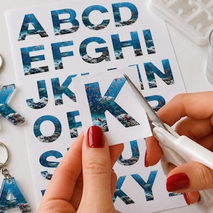 Könnte beinhalten: Ein weißes Blatt Papier, bedruckt mit dem Alphabet in einem blauen Unterwasser-Korallenriff-Muster. Eine weiße Schere schneidet den Buchstaben K aus. Zwei fertige Harz-Schlüsselanhänger in Form der Buchstaben K und A sind links zu sehen.