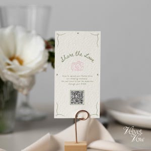 Può includere: Una carta rettangolare color bianco sporco con superficie testurizzata tenuta da un supporto in filo metallico color rame. La carta presenta angoli decorativi verdi, il testo "Share the Love", un'icona fotocamera rosa, un QR code e istruzioni.