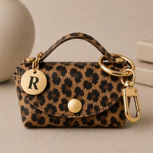 Puede incluir: Un peque&ntilde;o estuche con estampado de leopardo y asa superior, con cierre de bot&oacute;n a presi&oacute;n dorado, un mosquet&oacute;n dorado y un dije circular dorado grabado con la letra R. El estuche tiene una forma estructurada y herrajes en tono dorado.