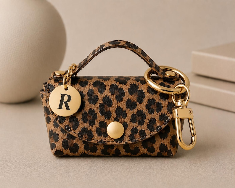 Puede incluir: Un peque&ntilde;o estuche con estampado de leopardo y asa superior, con cierre de bot&oacute;n a presi&oacute;n dorado, un mosquet&oacute;n dorado y un dije circular dorado grabado con la letra R. El estuche tiene una forma estructurada y herrajes en tono dorado.