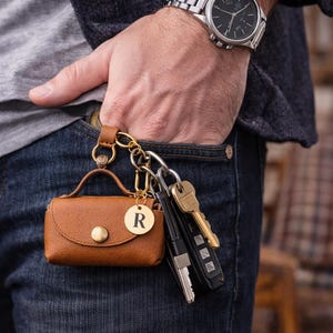 Puede incluir: Una peque&ntilde;a funda marr&oacute;n de aspecto cuero con asa superior y cierre de presi&oacute;n dorado, sujeta a un llavero dorado con un dije circular que muestra la letra R. La funda cuelga junto a un juego de llaves de metal y un mando de coche negro.