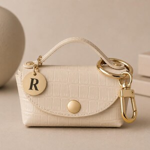 Puede incluir: Un mini bolso de color crema con relieve de cocodrilo, asa superior y cierre de bot&oacute;n a presi&oacute;n dorado. Incluye un llavero dorado con mosquet&oacute;n y un dije circular dorado grabado con la letra R en una fuente serif de color negro.