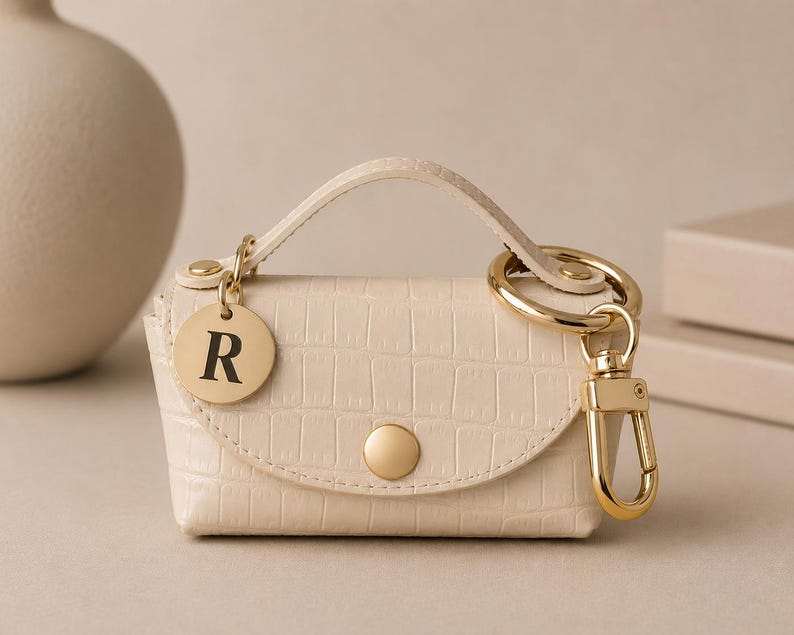 Puede incluir: Un mini bolso de color crema con relieve de cocodrilo, asa superior y cierre de bot&oacute;n a presi&oacute;n dorado. Incluye un llavero dorado con mosquet&oacute;n y un dije circular dorado grabado con la letra R en una fuente serif de color negro.