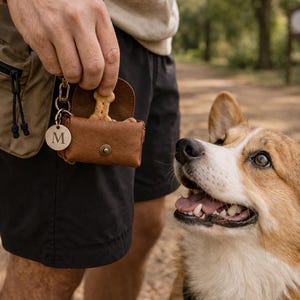 Puede incluir: Una peque&ntilde;a bolsa de cuero marr&oacute;n para premios con cierre de bot&oacute;n a presi&oacute;n, que contiene una golosina para perro en forma de hueso. La bolsa incluye un dije redondo de metal grabado con la letra M, unido a un clip dorado.