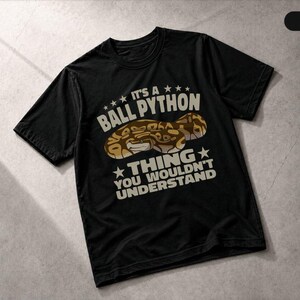 May include: A black short-sleeved t-shirt featuring a graphic of a brown and tan ball python. The text reads IT'S A BALL PYTHON THING YOU WOULDN'T UNDERSTAND in a light tan font, arranged above and below the snake illustration with star accents.