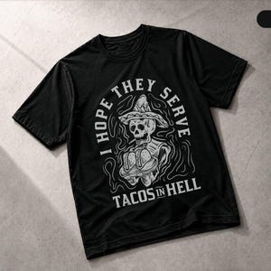 May include: A black short-sleeved t-shirt featuring a white graphic of a skeleton wearing a sombrero and holding two tacos. The text "I HOPE THEY SERVE TACOS IN HELL" is printed in white capital letters surrounding the central skeleton illustration.