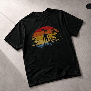 May include: A black short-sleeved t-shirt featuring a graphic print of a tall, thin creature with two speakers for a head. The design is set against a circular, multi-coloured sunset background with horizontal stripes in red, orange, and yellow.