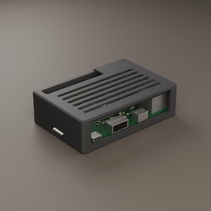 May include: A rectangular dark grey protective case for a single-board computer, featuring a vented top panel with parallel slats and an open side exposing the green circuit board, metallic ports, and various electronic components inside.