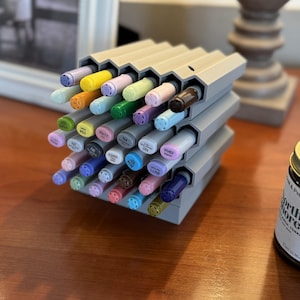 May include: A gray, modular, honeycomb-shaped plastic organizer holding a collection of multi-colored markers. The markers are arranged in individual hexagonal slots, displaying various pastel and vibrant cap colors with printed labels on each end.