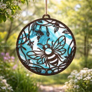 May include: Round wooden hanging decoration featuring a bee and floral design. The bee is in the center, surrounded by leaves and flowers. The background has a blue and white swirling pattern. The decoration is suspended by a natural-colored string.