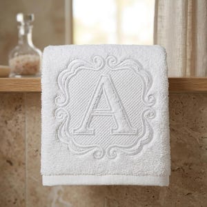 May include: A white terry cloth hand towel featuring a centered embroidered letter A. The letter is enclosed within a decorative, stitched frame with swirling scrollwork details, creating a textured, monochromatic design on the fabric.
