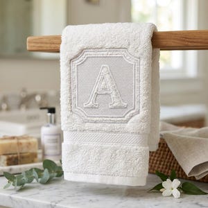 Può includere: Un asciugamano in cotone bianco con un riquadro quadrato ricamato in grigio chiaro al centro, caratterizzato da un bordo decorativo e dalla lettera maiuscola A. L'asciugamano ha una finitura in spugna testurizzata ed è appeso a una barra di legno.
