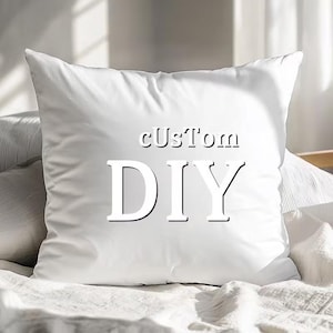 Op de afbeelding: Een vierkant wit sierkussen met de tekst cUsTom DIY in een vetgedrukt, donker lettertype in het midden van de voorzijde. Het kussen heeft een gladde, effen witte stoffen afwerking en staat rechtop, wat zorgt voor een strak en eenvoudig ontwerp.