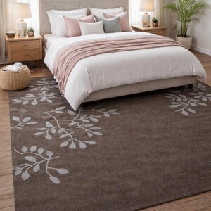 May include: A bedroom scene featuring a bed with white bedding, pink and gray pillows, and a blush throw. A brown rug with a white leaf design is in the foreground. Two wooden nightstands with lamps flank the bed. A potted plant is in the background.