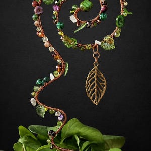 May include: A copper wire plant trellis featuring a spiral top and a wavy stem, decorated with various green, brown, and clear beads, small glass leaves, and a single dangling metal leaf charm, positioned upright in a green leafy houseplant.