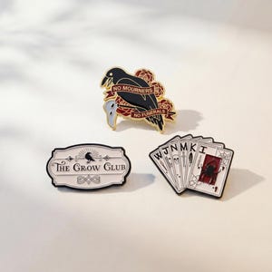 May include: Three enamel pins featuring a black crow with red roses and the text NO MOURNERS NO FUNERALS, a white rectangular sign with the text THE CROW CLUB and a crow silhouette, and a set of playing cards with a spider and various symbols.