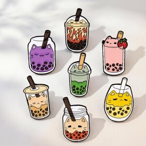 May include: A collection of seven enamel pins shaped like bubble tea cups, each featuring a cat face design. The pins display various colours including purple, pink, green, yellow, and brown, with black straws and dark boba pearls inside.