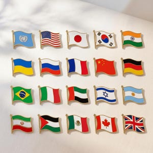 May include: A collection of twenty rectangular metal lapel pins shaped like waving flags. The set includes various national flags and the United Nations emblem, each featuring vibrant enamel colours and gold-toned metal edges arranged in four rows.