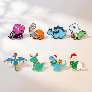 May include: A set of eight enamel pins featuring cartoon dinosaurs in various costumes. The top row includes a pink triceratops skeleton, an orange mummy, a blue stegosaurus, and a green ghost holding a pumpkin. The bottom row shows a blue dragon, a green reindeer, a blue dinosaur with a gift, and a white dinosaur in a green sweater, all wearing holiday hats.