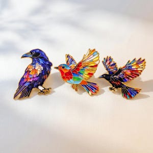 Puede incluir: Tres pines de esmalte metálico con diseños de aves y patrones estilo vitral. El pin de la izquierda es un cuervo posado, el central es un petirrojo en vuelo con colores vibrantes y el de la derecha es un ave negra volando con alas multicolores.