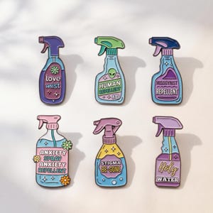 Könnte beinhalten: Sechs Emaille-Anstecknadeln in Form von Sprühflaschen in verschiedenen Farben. Die Anstecknadeln tragen Aufschriften wie love mist, human repellent, misogynist repellent, anxiety spray anxiety repellent, stigma be-gone und holy water, verziert mit kleinen Sternen und Blumen.