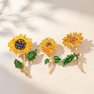 Op de afbeelding: Drie zonnebloem broches van goudkleurig metaal met groene emaille bladeren. De linker broche heeft gele bloemblaadjes en een centrum van donkere iriserende kristallen. De middelste en rechter broches hebben getextureerde goudkleurige centra.