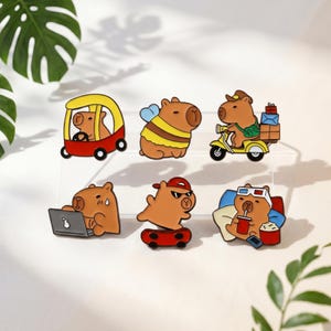 Puede incluir: Un conjunto de seis pines de esmalte con capibaras marrones en diversas actividades: conduciendo un coche rojo, disfrazado de abeja, en un scooter con paquetes, trabajando en un portátil, andando en monopatín y viendo una película con gafas 3D.