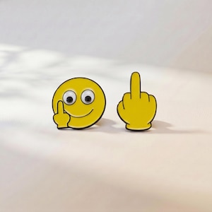 May include: Two yellow enamel pins. One pin is a circular smiley face with googly eyes and a hand making a middle finger gesture. The second pin is a yellow hand shape with a raised middle finger, featuring black outlines and a cartoon style.