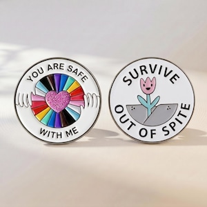 May include: Two circular enamel pins. The left pin features a glittery pink heart surrounded by a rainbow sunburst, white hands, and the text "YOU ARE SAFE WITH ME". The right pin shows a pink flower in grey soil with the text "SURVIVE OUT OF SPITE".