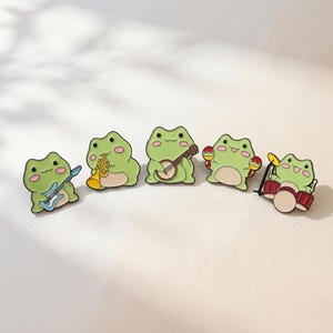 May include: A set of five enamel pins featuring light green frogs, each playing a different musical instrument: a blue electric guitar, a yellow trumpet, a brown banjo, red maracas, and a red drum set with a yellow cymbal.