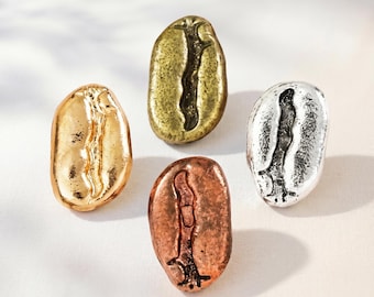 Könnte beinhalten: Vier Metallanstecker in Form von Kaffeebohnen, jede mit einem anderen Finish. Eine ist aus poliertem Gold, eine aus antikem Messing, eine aus Kupfer und eine aus Silber. Jede Bohne hat eine zentrale vertikale Rille und eine strukturierte Oberfläche.