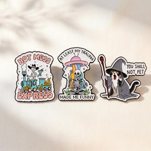Pode incluir: Três pins de esmalte com ilustrações coloridas. O primeiro mostra um esqueleto num comboio com o texto "HOT MESS EXPRESS". O segundo representa um esqueleto sob um OVNI com "AT LEAST MY TRAUMA MADE ME FUNNY". O terceiro mostra um gato com um chapéu de feiticeiro e "YOU SHALL NOT PET".