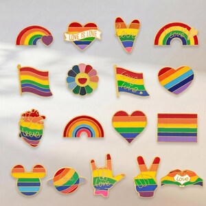 Puede incluir: Una colección de dieciocho pines de esmalte con diseños de colores del arcoíris que incluyen corazones, banderas, una flor, un órgano anatómico en forma de corazón, un planeta, una silueta de Mickey Mouse, gestos con las manos y el texto love is love.