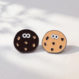 Pode incluir: Dois pins redondos de esmalte em forma de biscoito com rostos de desenho animado. Um pin é castanho escuro com pedaços bege, e o outro é bege com pedaços castanho escuro. Ambos têm olhos brancos grandes e uma boca pequena.