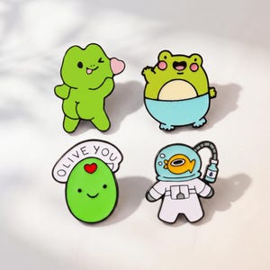 May include: Four enamel pins featuring cartoon characters: a green frog winking, a green frog in blue shorts, a green olive with a red heart and the text OLIVE YOU, and a fish in a white astronaut suit with a blue oxygen tank and tube.