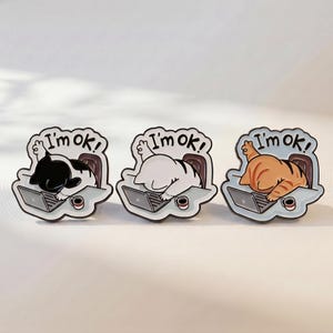Op de afbeelding: Drie emaille pins met een zwart-witte kat, een witte kat en een oranje cyperse kat, elk voorovergebogen over een laptop met een koffiemok. De tekst "I'm ok!" staat in het zwart boven elke kat afgebeeld in een grappige werksituatie.