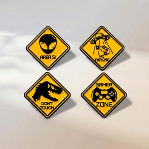 Peut inclure: Quatre broderies en émail en forme de losange avec des fonds jaunes et des bordures noires. La première montre une tête d'alien et le texte AREA 51. La deuxième montre un OVNI soulevant une voiture et le texte NO PARKING. La troisième montre un dinosaure et le texte DON'T TOUCH. La quatrième montre une manette de jeu et le texte GAMER ZONE.