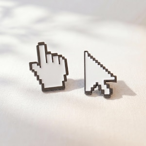May include: A pair of enamel pins featuring pixelated computer interface icons. One pin is a white hand cursor with a black outline, and the other is a white arrow pointer with a black outline, both designed with a distinct blocky aesthetic.