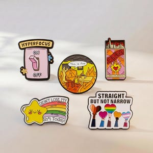 Könnte beinhalten: Fünf Emaille-Anstecknadeln mit verschiedenen Motiven: ein Lichtschalter mit der Aufschrift HYPERFOCUS, ein runder Pin mit einem Würfel im Feuer und dem Text This is Fine, eine Streichholzschachtel mit SLOW BURN, ein Stern mit Regenbogen und Text sowie ein Schild mit STRAIGHT BUT NOT NARROW.