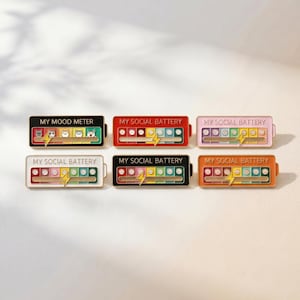 May include: Six rectangular enamel pins shaped like batteries, each featuring a sliding lightning bolt icon and a row of emoji faces. The pins are labeled with the text MY MOOD METER or MY SOCIAL BATTERY and come in black, red, pink, white, and orange.