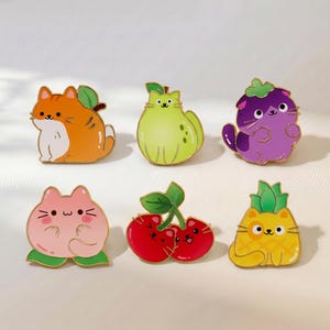 May include: A set of six enamel pins featuring cartoon cats shaped like various fruits. The collection includes an orange cat shaped like a peach, a light green cat pear, a purple cat eggplant, a pink cat peach, two red cherry cats, and a yellow pineapple cat.