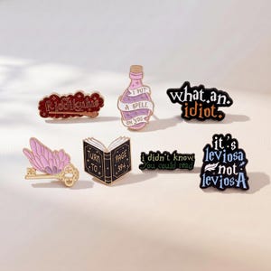 May include: A collection of seven enamel pins featuring various designs including a red glitter Riddikulus sign, a pink potion bottle, a black what an idiot sign, a winged key, an open book, a green sign, and a blue it's leviosa not leviosa sign.