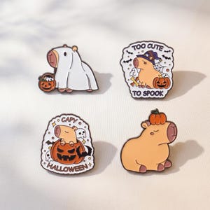 Puede incluir: Cuatro pines de esmalte con capibaras con temática de Halloween. Uno muestra un capibara disfrazado de fantasma con un cubo de calabaza, otro dice TOO CUTE TO SPOOK con un sombrero de bruja, uno dice CAPY HALLOWEEN con una calabaza y otro tiene una.
