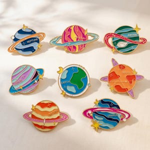 Könnte beinhalten: Ein Set aus acht bunten Emaille-Anstecknadeln in Planetenform, darunter Neptun, Merkur, Jupiter, Venus, Erde, Mars, Saturn und Uranus. Jede Anstecknadel zeichnet sich durch eine eigene Farbpalette, metallische Umrandungen und kleine Sterne aus.