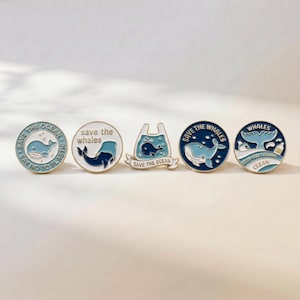 May include: Five circular and shaped enamel pins featuring blue whales and ocean conservation themes. The pins include text such as SAVE THE OCEAN, SAVE THE WHALES, and KEEP OUR OCEANS CLEAN, with designs depicting whales in water, plastic bags, and bubbles.